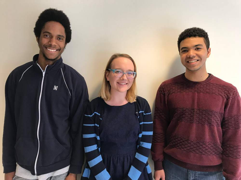 Three people stand against a gray wall. They are two boys and a girl in the middle.